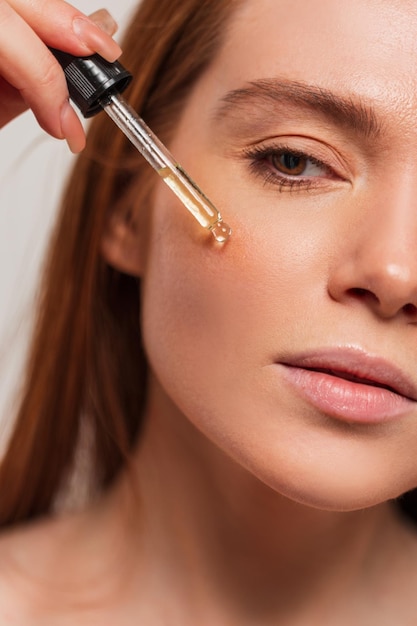 Belle jeune femme fraîche aux cheveux roux aux yeux bruns applique de l'huile sur son visage avec une pipette Beauté soins de la peau et hydratation