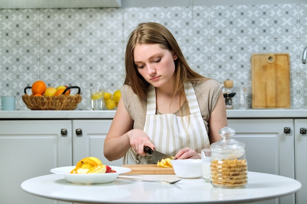 Belle jeune femme avec des fruits dans la cuisine, fille assise à table et coupe l'orange. Blogueuse culinaire cuisine salade de fruits à la caméra