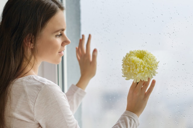 Belle jeune femme avec une grande fleur jaune pâle près de la fenêtre