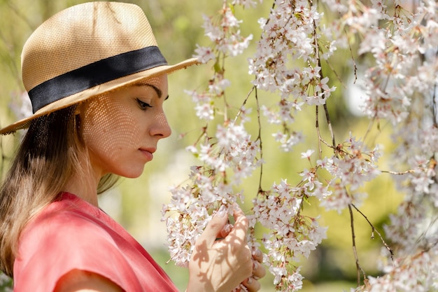 Une belle jeune femme en gros plan dans un chapeau de paille admire les fleurs de cerisier blanches et lumineuses dans le jardin de printemps