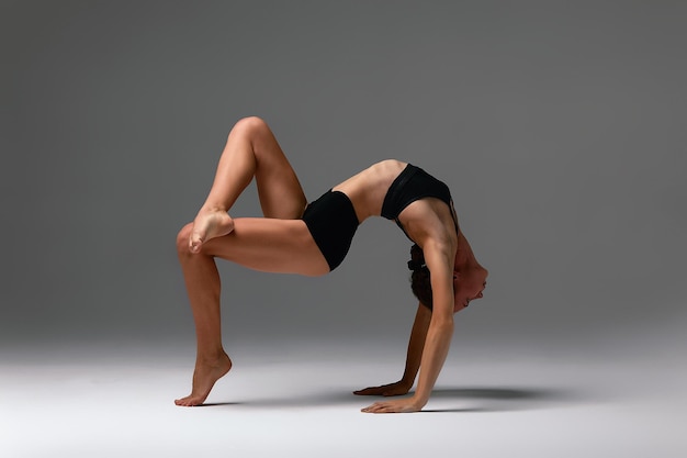 Belle jeune femme gymnaste flexible en justaucorps noir travaillant sur des étirements faisant de l'exercice de backbend pleine longueur studio fond gris