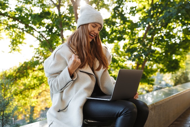 Belle jeune femme habillée en manteau d'automne et chapeau assis à l'extérieur, à l'aide d'un ordinateur portable, célébrant le succès