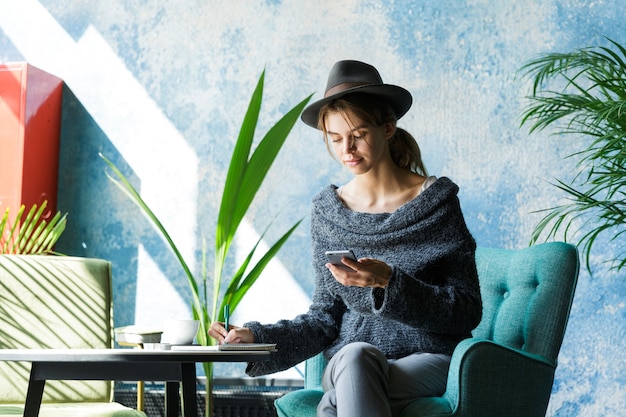 Belle jeune femme habillée en pull et chapeau assis dans une chaise à la table du café, à l'aide de téléphone portable, prendre des notes, intérieur élégant