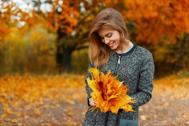 Belle jeune femme heureuse dans des vêtements d'automne à la mode tient des feuilles jaunes à l'extérieur