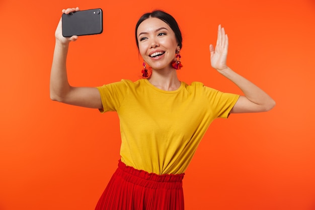 belle jeune femme heureuse posant isolée sur un mur orange parlant par téléphone portable prendre un selfie en agitant.