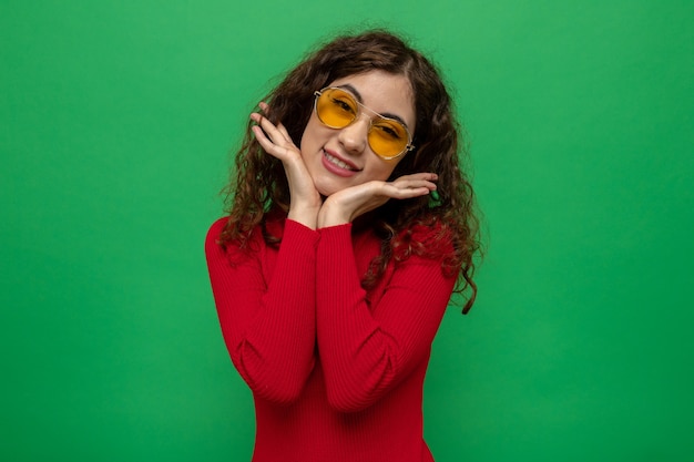 Belle jeune femme heureuse et positive en col roulé rouge portant des lunettes jaunes souriant joyeusement avec les mains sur son visage debout sur un mur vert