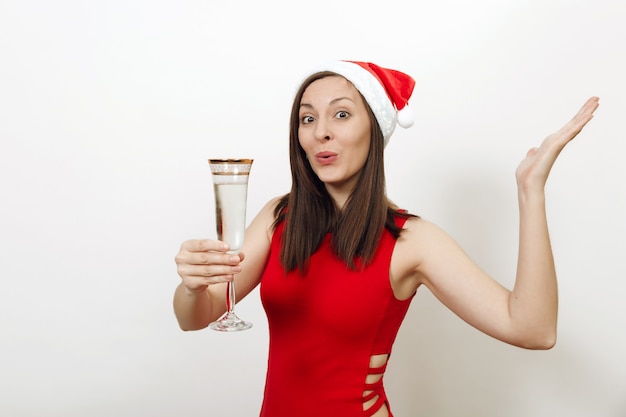 Belle jeune femme heureuse de race blanche avec un charmant sourire en robe rouge et chapeau de Noël debout avec une coupe de champagne sur fond blanc. Fille de Santa isolée. Concept de vacances nouvel an 2018.