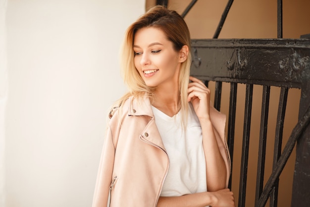 Belle jeune femme heureuse avec un sourire dans une veste rose élégante près d'une clôture métallique dans la rue