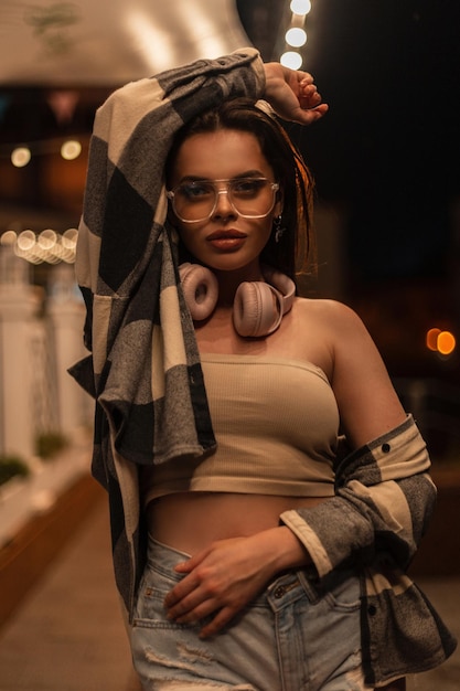 Belle jeune femme hipster élégante avec des lunettes de mode et des écouteurs dans des vêtements décontractés à la mode avec un haut et une chemise à carreaux se dresse dans la ville de nuit