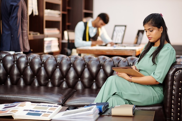 Belle jeune femme indienne assise sur un canapé en atelier et regarder le catalogue sur ordinateur tablette