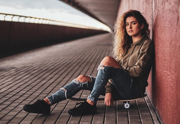 Belle jeune femme en jeans déchirés est assise sur son propre skateboard dans la rue.