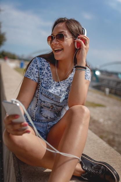 Une belle jeune femme joyeuse écoutant de la musique depuis votre téléphone et profitant de la rive de la ville par une belle journée d'été.