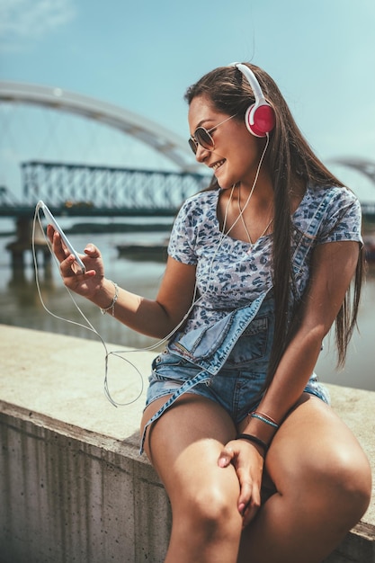 Une belle jeune femme joyeuse écoute la musique de son smartphone et profite de la rive de la rivière de la ville par une belle journée d'été.