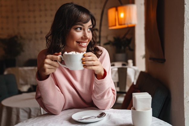 une belle jeune femme joyeuse et optimiste à l'intérieur dans un café buvant du café avec de la guimauve.