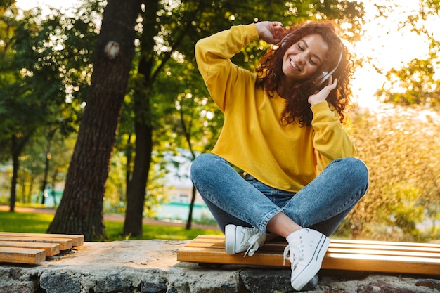 Belle jeune femme joyeuse passant du bon temps au parc, assise sur un banc, écoutant de la musique avec des écouteurs