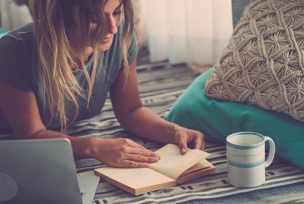 Belle jeune femme lisant un livre en position allongée sur un tapis à côté d'un ordinateur portable et d'un verre à la maison. Femme passant son temps libre allongée sur un tapis et lisant un livre à la maison