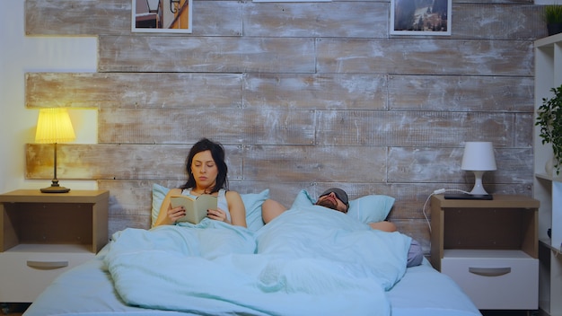 Belle Jeune Femme Lisant Un Livre En Pyjama Pendant Que Le Mari Couvre Le Visage Avec Un Masque De Sommeil.