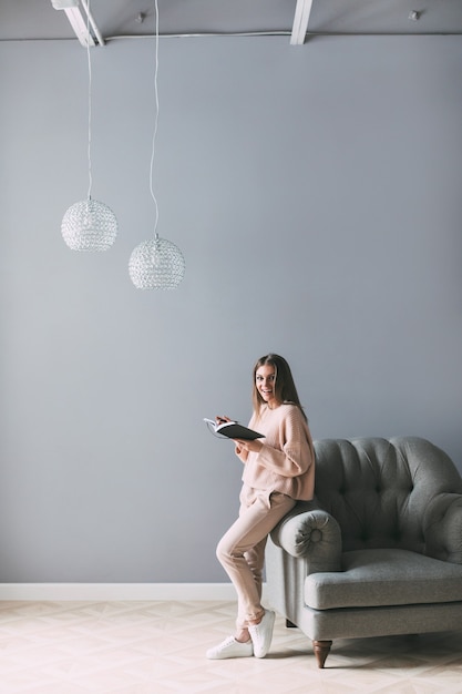 Belle jeune femme avec un livre près de la chaise à la maison. Écrire dans le journal.