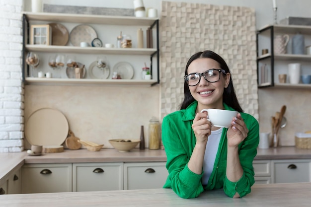 Belle jeune femme à lunettes et une chemise décontractée verte à la maison dans la cuisine regarde la webcam