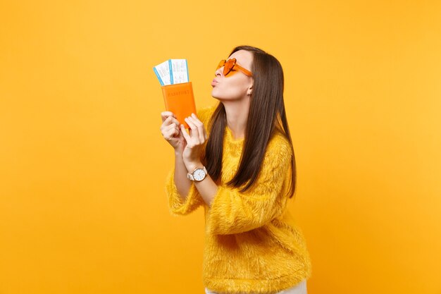 Belle jeune femme à lunettes coeur orange soufflant des lèvres envoyant un baiser aérien tenant un passeport, des billets d'embarquement isolés sur fond jaune. Les gens émotions sincères, mode de vie. Espace publicitaire.