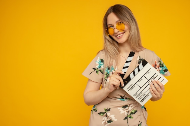 Belle jeune femme à lunettes orange tenant un film blanc classique faisant un clap isolé sur fond jaune. Les gens émotions sincères, concept de style de vie. Espace publicitaire