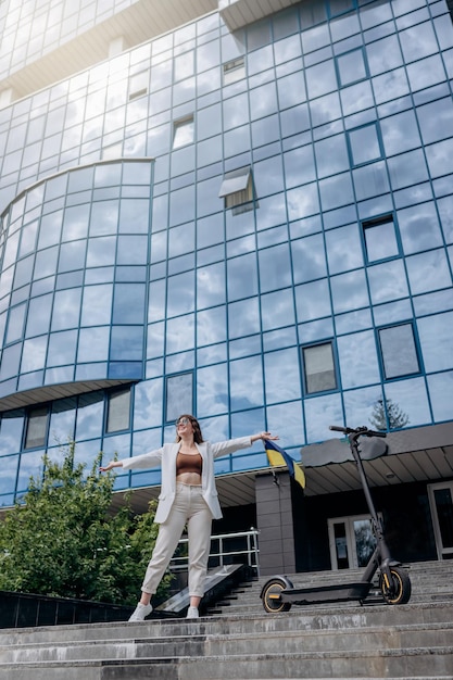 Belle jeune femme en lunettes de soleil et costume blanc debout avec son scooter électrique près d'un bâtiment moderne et regardant loin