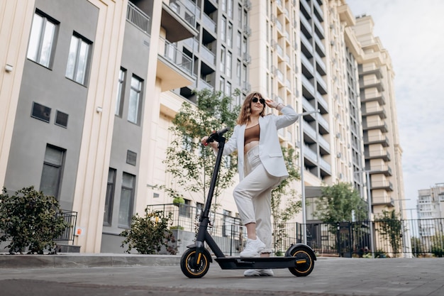 Belle jeune femme en lunettes de soleil et costume blanc debout sur son scooter électrique près d'un bâtiment moderne et regardant loin