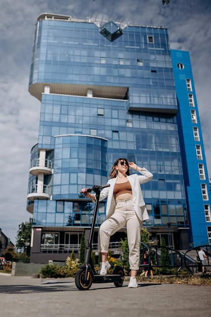 Belle jeune femme en lunettes de soleil et costume blanc debout sur son scooter électrique près d'un bâtiment moderne et regardant loin