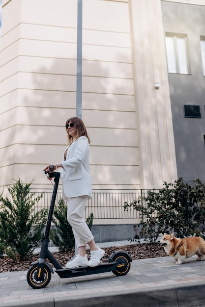 Belle jeune femme en lunettes de soleil et costume blanc monte sur son scooter électrique avec un chien Corgi près d'un bâtiment moderne en ville et regarde au loin