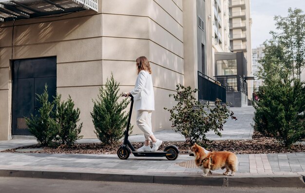 Belle jeune femme en lunettes de soleil et costume blanc monte sur son scooter électrique avec un chien Corgi près d'un bâtiment moderne en ville et regarde au loin