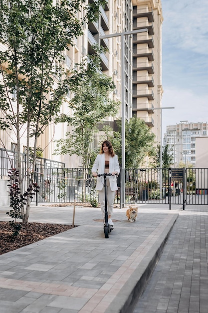 Belle jeune femme en lunettes de soleil et costume blanc monte sur son scooter électrique avec un chien Corgi près d'un bâtiment moderne en ville et regarde au loin