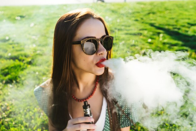 Belle Jeune Femme à Lunettes De Soleil Est Assise Sur La Pelouse Avec De L'herbe Verte Brillante, Fumant, Regardant Vers La Droite. Journée Ensoleillée