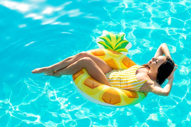 Belle jeune femme en maillot de bain relaxante sur ananas jaune en forme d'anneau en caoutchouc gonflable dans la piscine. Profiter de l'été. Ambiance vacances.