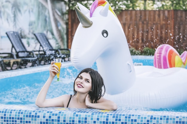 Belle jeune femme en maillot de bain rose dans la piscine lors d'un cocktail avec une licorne gonflable