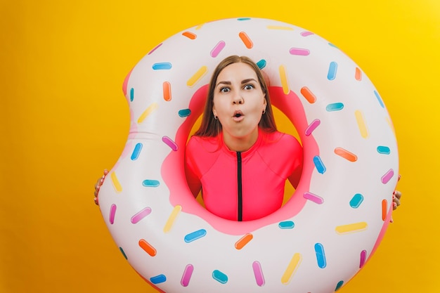 Belle jeune femme en maillot de bain rose élégant avec un anneau gonflable beignet sur fond jaune