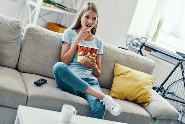 Belle jeune femme mangeant du pop-corn en regardant la télévision sur le canapé à la maison