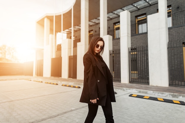 Belle jeune femme mannequin moderne en robe noire décontractée à la mode dans des lunettes de soleil élégantes parcourt la rue au coucher du soleil. Belle fille à la mode sur fond de soleil orange à l'extérieur. Regard de printemps.