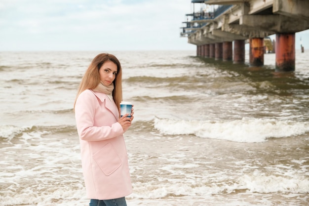 Une belle jeune femme en manteau rose se tient au bord de la mer ou de l'océan avec une tasse de café