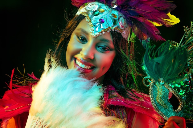 Belle jeune femme en masque de carnaval et costume de mascarade élégant avec ventilateur de plumes dans des lumières colorées et lueur sur fond noir