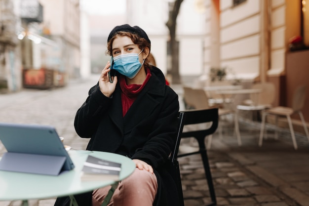 Belle jeune femme en masque de protection parlant sur mobile alors qu'il était assis à l'extérieur sur la terrasse du café. Concept de maladie infectieuse et de temps de pandémie.