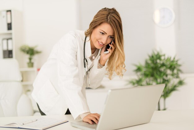Belle jeune femme médecin dans sa salle de consultation, utilisant le téléphone et écrivant sur un ordinateur portable.