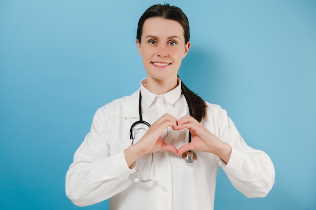 Belle jeune femme médecin montrant un geste cardiaque et souriant, prenant soin des patients avec amour, porte un stéthoscope de blouse de laboratoire blanc, des modèles sur un mur de fond bleu studio. Covid-19, concept de pandémie