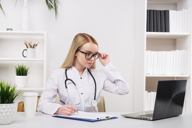 Belle jeune femme médecin travaillant heureux et sourire à l'hôpital, assis sur la table, concept médical