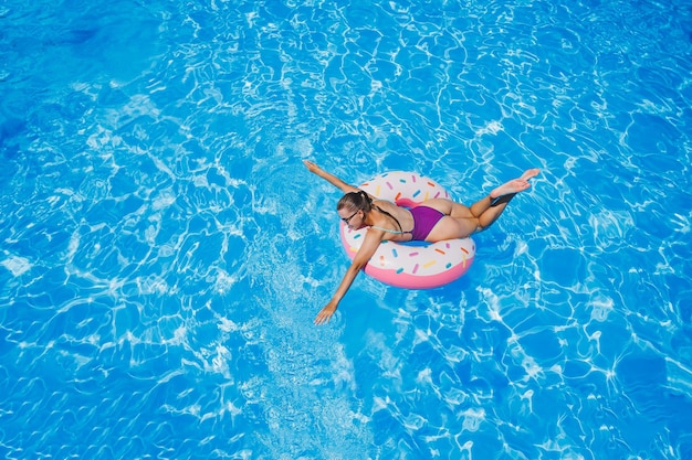 Belle jeune femme mince en maillot de bain profitant du parc aquatique flottant dans un grand anneau gonflable sur une piscine bleue étincelante souriant à la caméra Vacances d'été