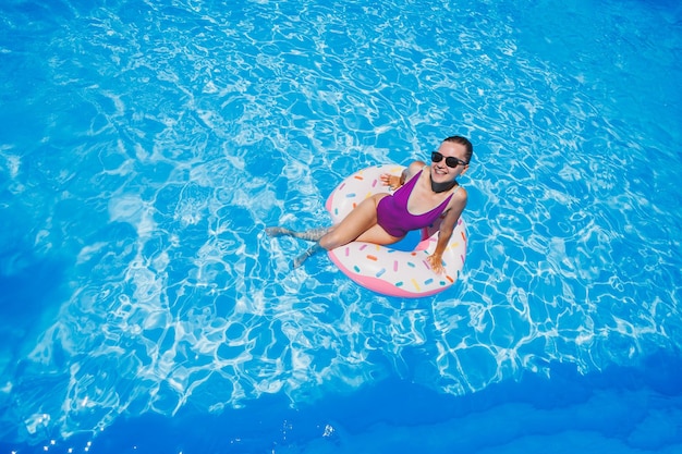 Belle jeune femme mince en maillot de bain profitant du parc aquatique flottant dans un grand anneau gonflable sur une piscine bleue étincelante souriant à la caméra Vacances d'été