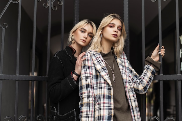 Belle jeune femme à la mode en blazer noir posant avec sa petite amie dans une élégante veste grise à carreaux près de la porte de fer