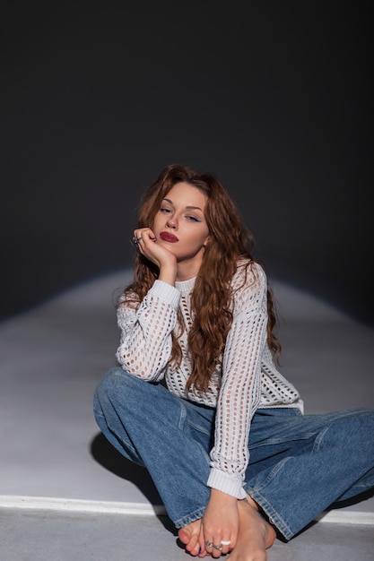 Une belle jeune femme à la mode dans un pull blanc à la mode avec des jeans vintage est assise sur le sol dans le studio.