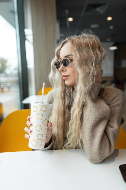 Belle jeune femme modèle hipster avec une coiffure blonde bouclée avec des lunettes de soleil en tenue de mode se trouve dans un café et boit du cola