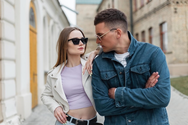 Belle jeune femme modèle et un homme hipster dans des vêtements en denim à la mode avec des lunettes de soleil se promènent dans la ville Couple urbain à la mode à l'extérieur