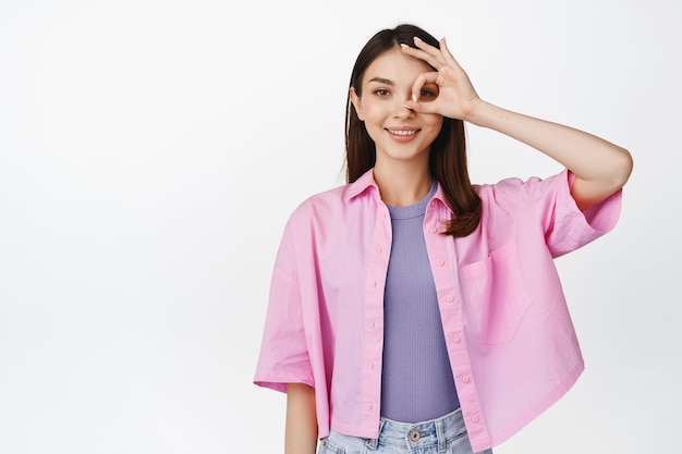 Belle jeune femme montre un geste correct zéro sur les yeux en souriant et en regardant la caméra confiant aucun signe de problème debout sur fond blanc